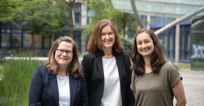 Drei professional gekleidete Frauen lächelnd vor einem Bürogebäude in grüner Umgebung